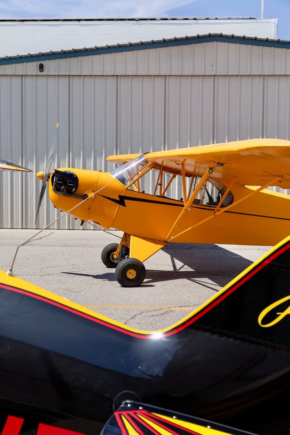 A yellow Piper J-3 Cub on the ramp at Van Nuys Airport, with the black-and-red wingtip of a Pitts S-2B biplane in the foreground